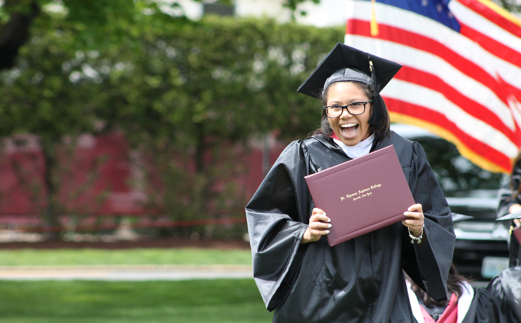 A female in a black graduation gown holding a burgundy diploma cover.