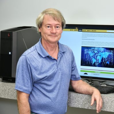 Dr. Vermilyer smiling wearing blue dress shirt next to computer in classroom