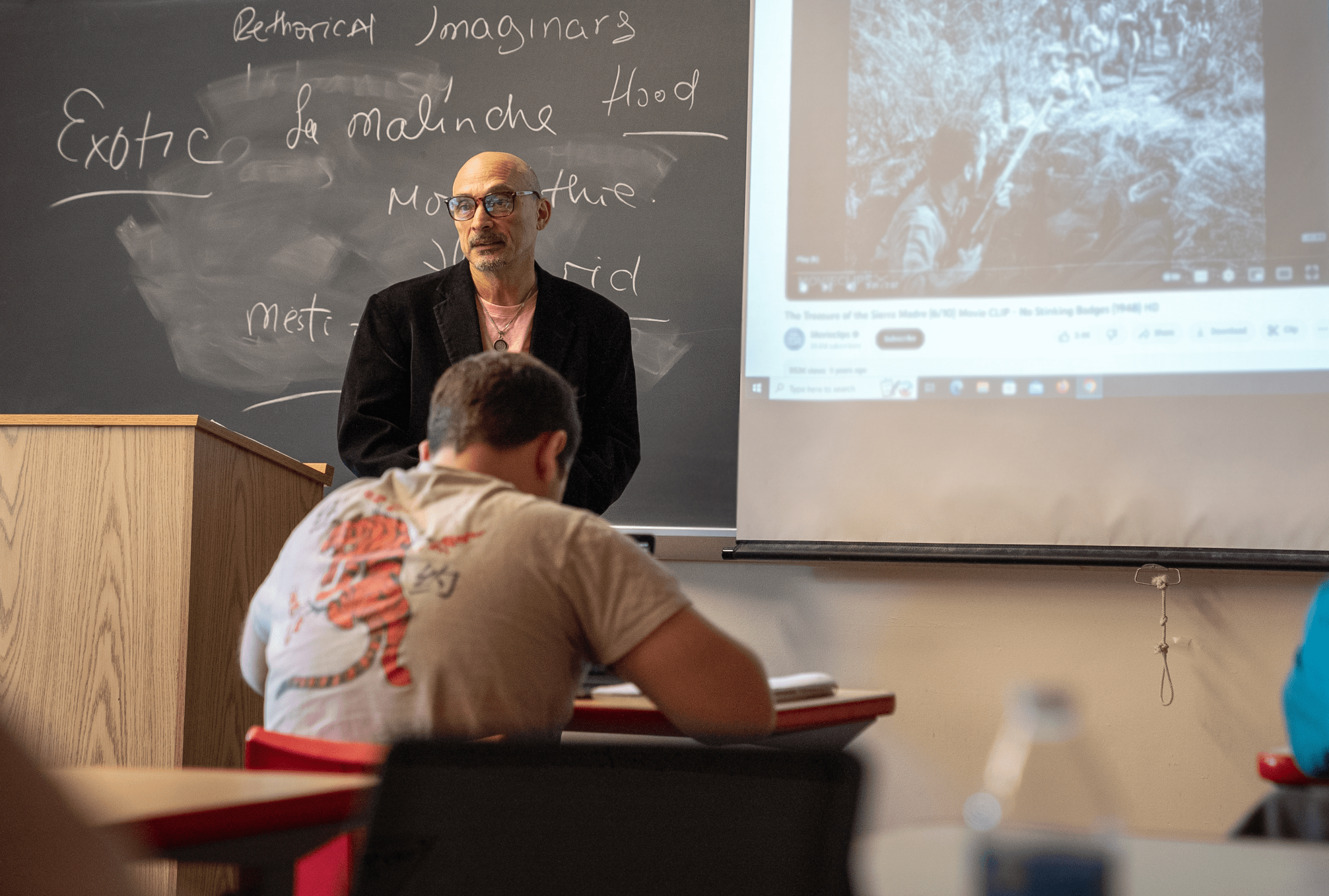 Professor Baretto teaching in classroom with screen showing black and white video in background