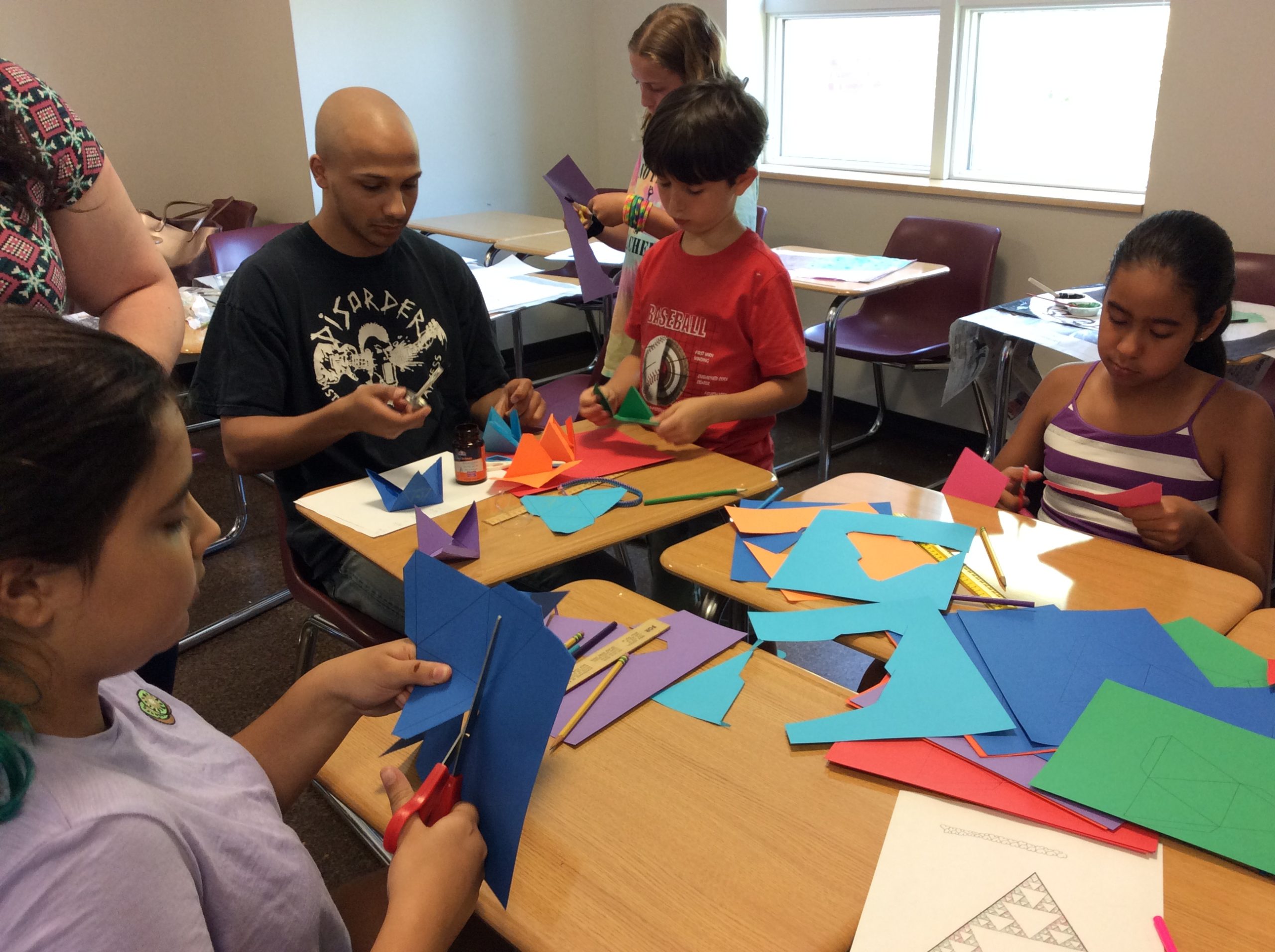 Students and children in a classroom sitting at desks and cutting shapes from papers of different color.