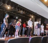 12 students on stage at the Sullivan theatre in regular street clothes practicing for a show, all looking forward towards audience chairs