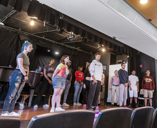 12 students on stage at the Sullivan theatre in regular street clothes practicing for a show, all looking forward towards audience chairs