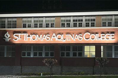 St. Thomas Aquinas College signage on Costello Hall building at night lit up with glowing white light.