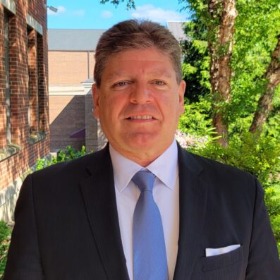 Mike Greco smiling wearing black suit and blue tie with green tree in background