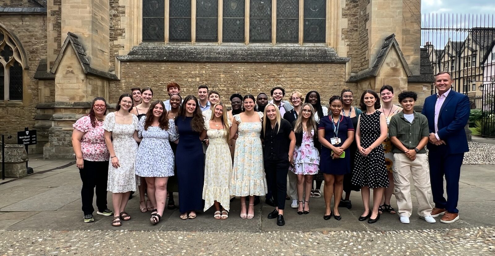 group of student studying in Oxford outside wearing formal attire smiling for photo. Oxford University building in background