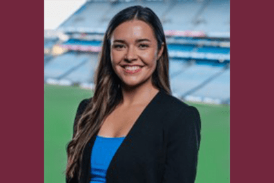 Mary Kate Henry smiling wearing black blazer and blue shirt with Stadium in background